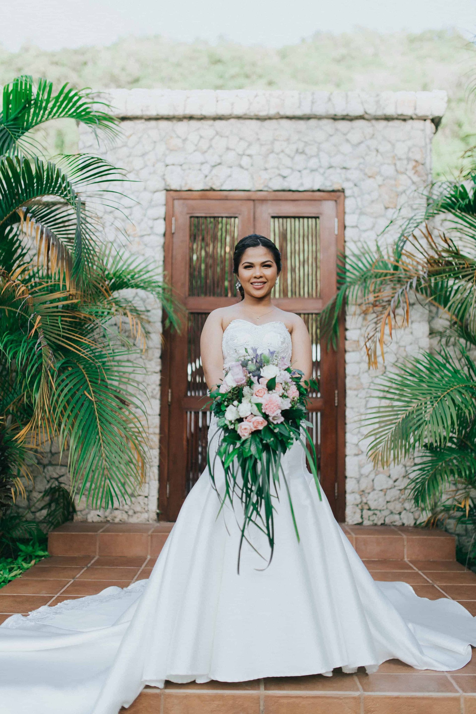 Bride in white gown holding bouquet, standing in front of a stone building, surrounded by greenery.
