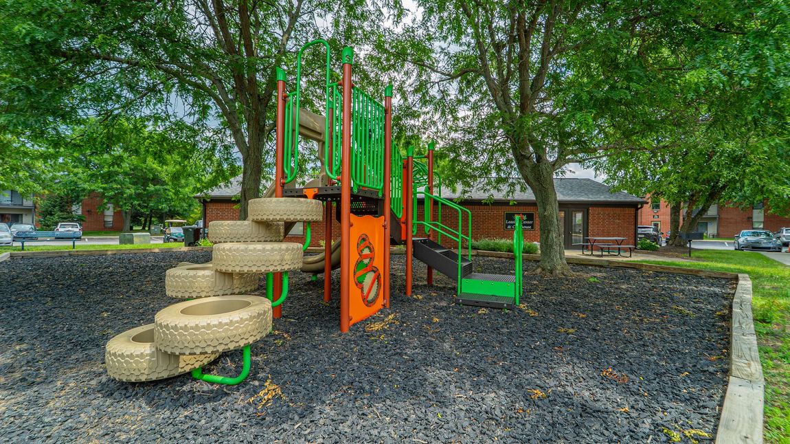 Playground with tire steps, slides, and green and orange structures, surrounded by trees and black mulch.