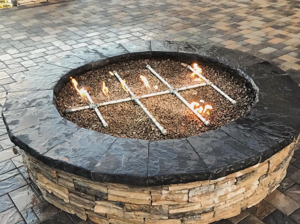 Circular stone fire pit with a metal grate and small flames burning inside on a paved courtyard.