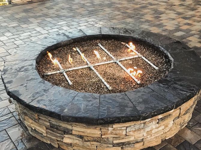 Circular stone fire pit with a metal grate and small flames burning inside on a paved courtyard.
