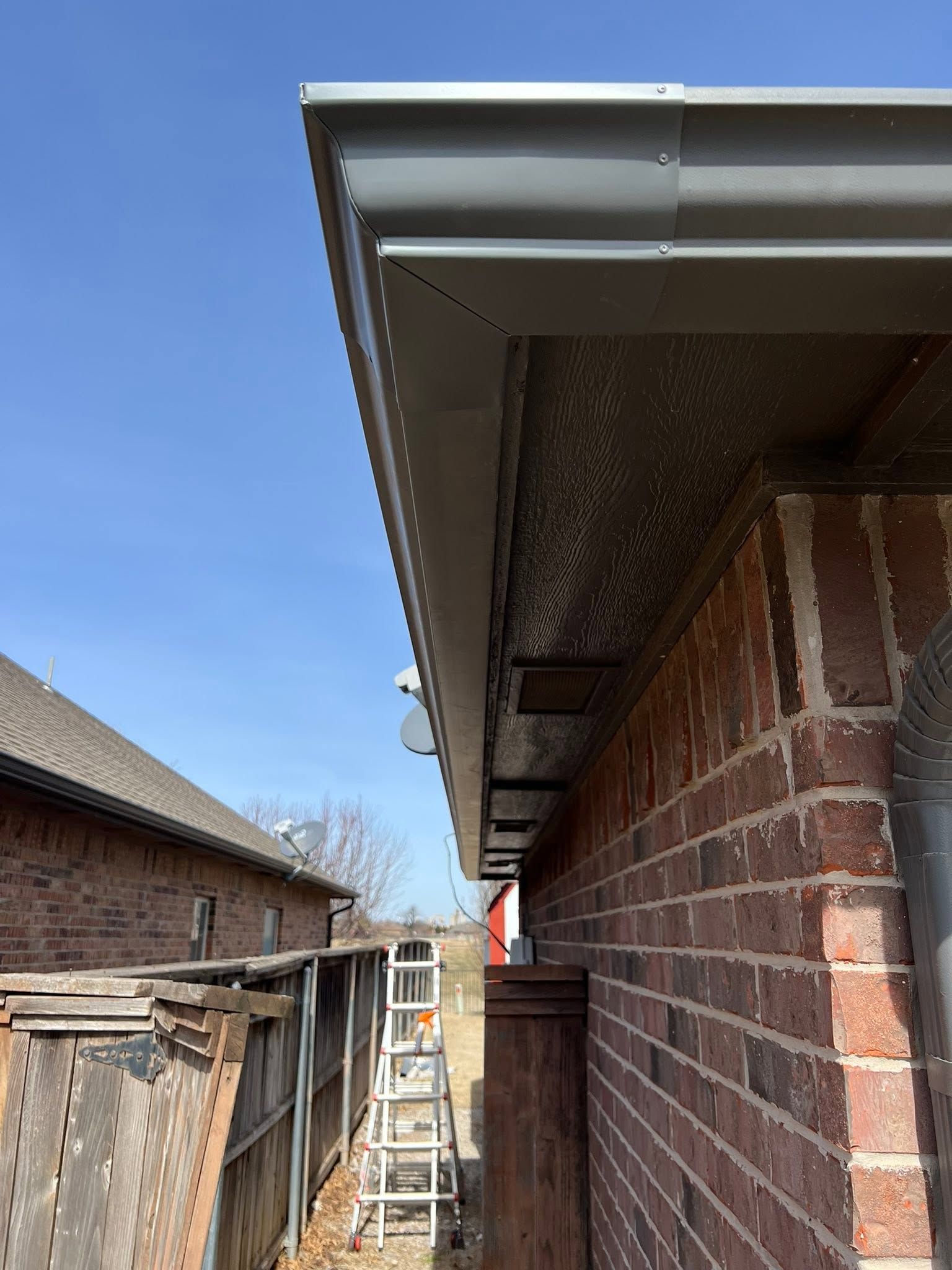 A ladder beside a brick house under the roof eaves, with a fence and blue sky in the background