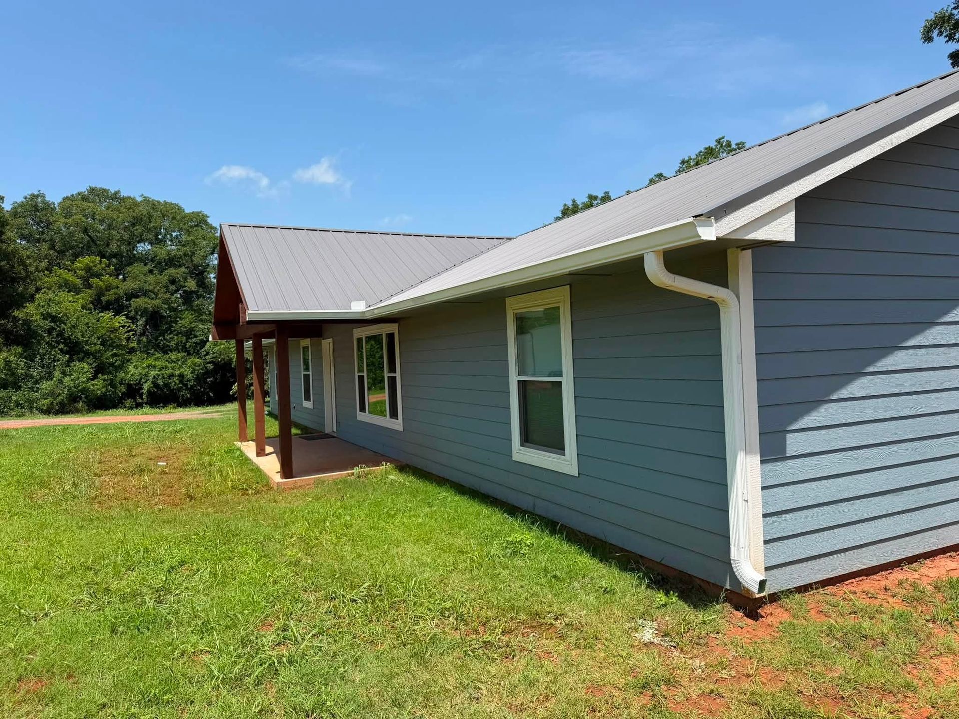 Single-story blue house with white trim and a metal roof on a grassy lot under a clear sky