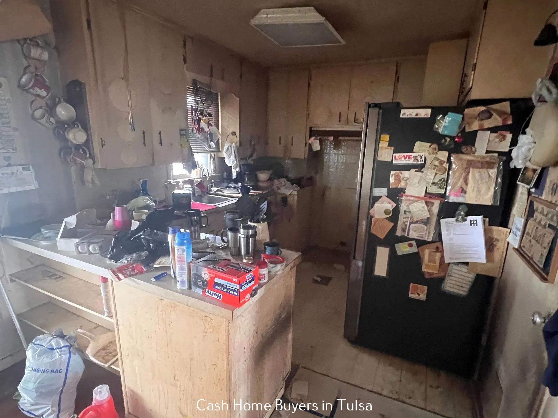 A cluttered, dated kitchen with worn wooden cabinets, a black refrigerator covered in notes, and a central workspace.
