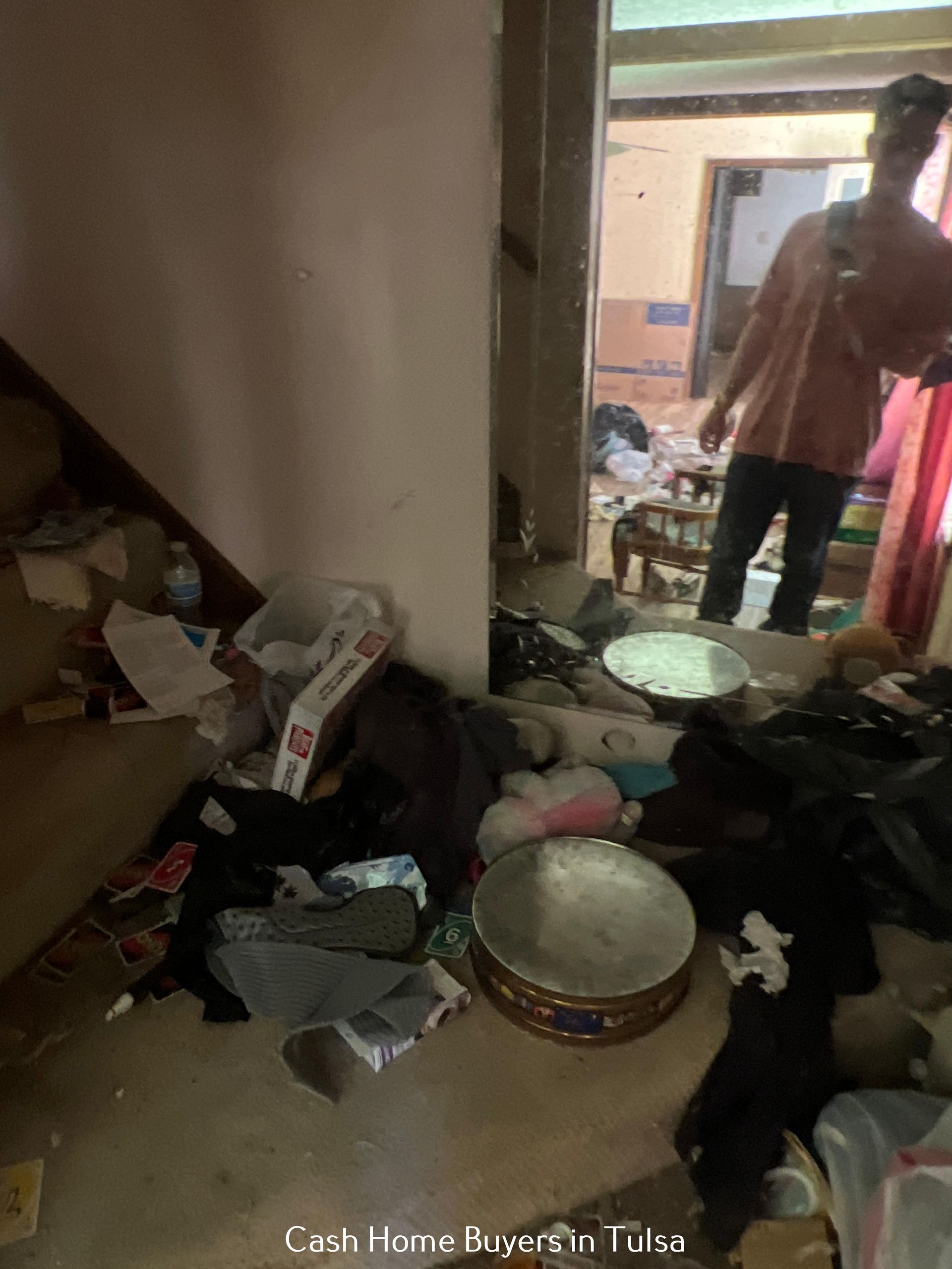 A person stands in a doorway, reflected in a large mirror, overlooking a cluttered, debris-filled room.