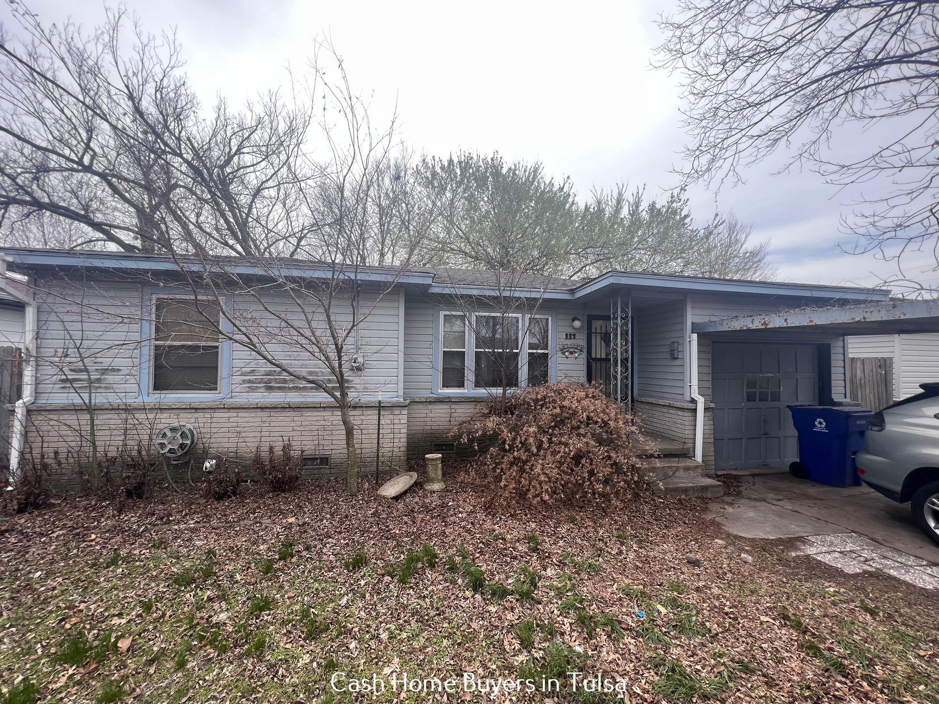 A light gray single-story house with a white stone base, a front yard covered in fallen leaves, and a side carport.