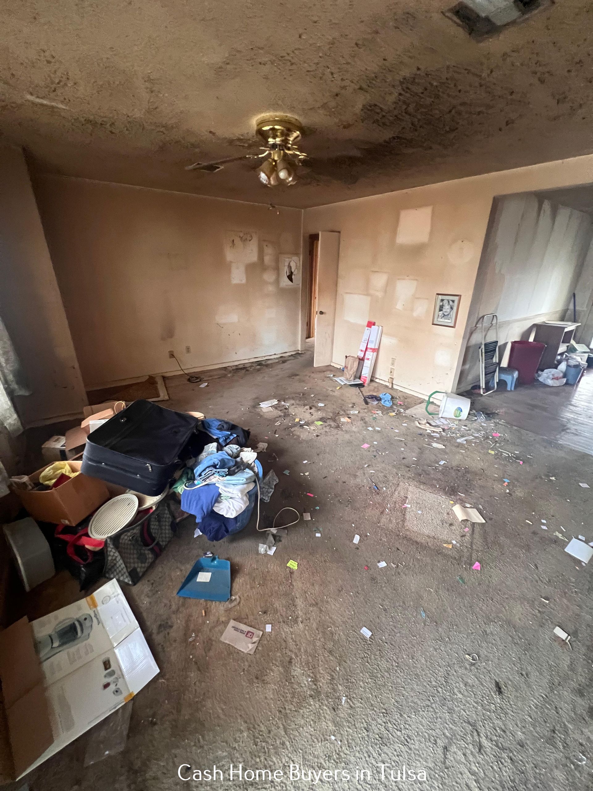 A cluttered, dilapidated room with debris on the floor, dark stains on the ceiling, and a ceiling fan.
