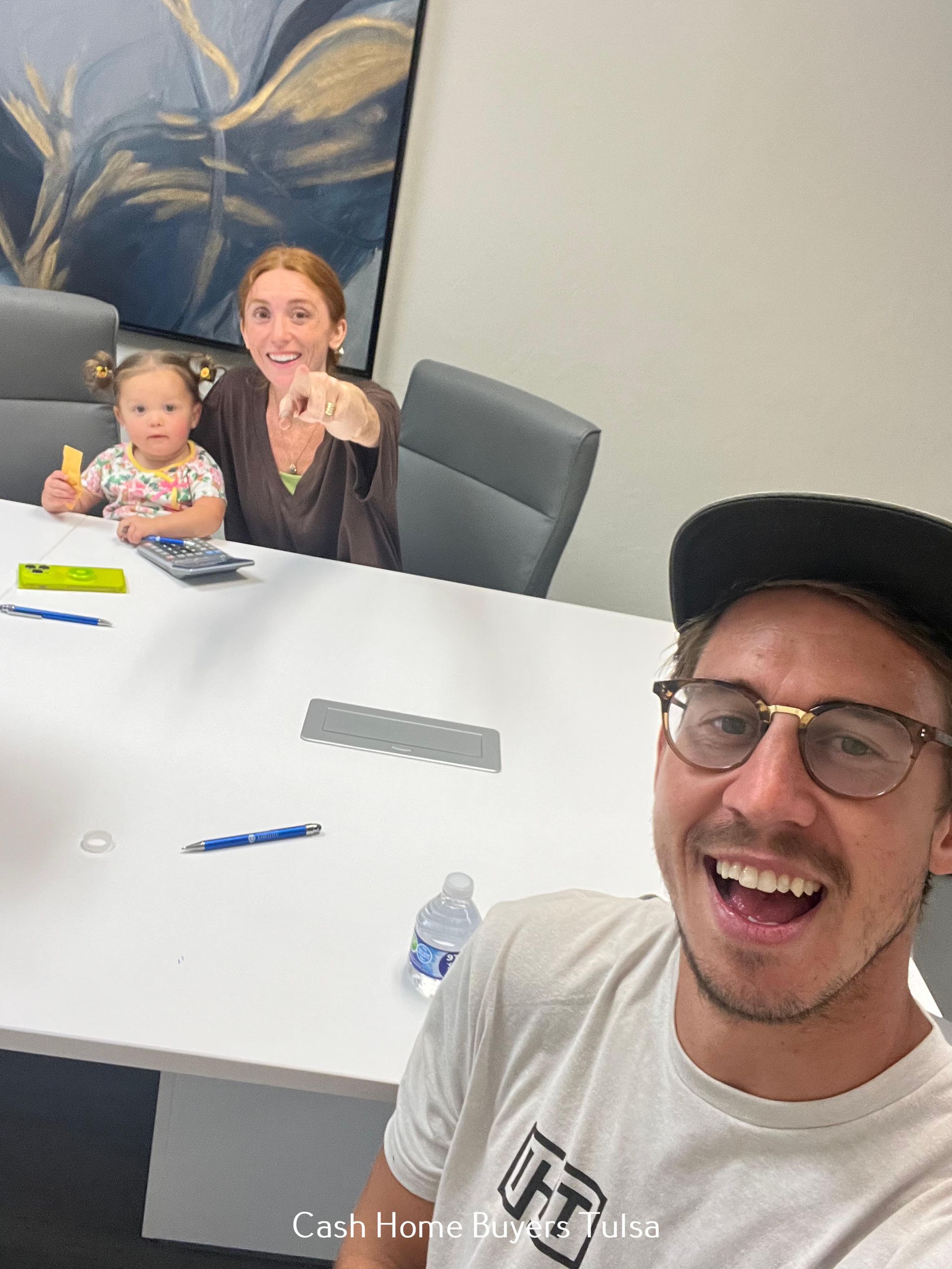 Two people and a baby at a conference table, with a smiling man taking a selfie in the foreground.