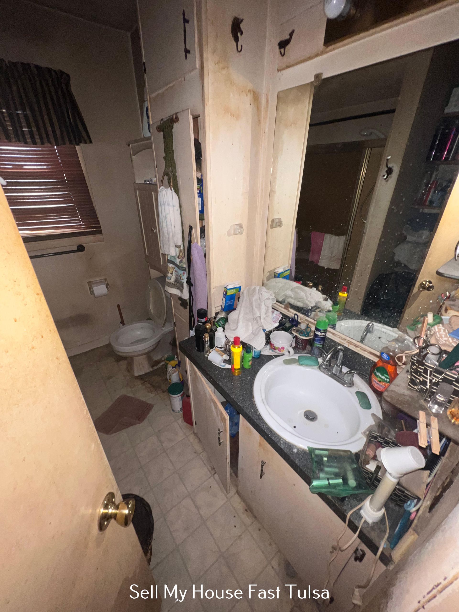 A cluttered bathroom with wood-look cabinets, a white sink, and a toilet, viewed from an open doorway.