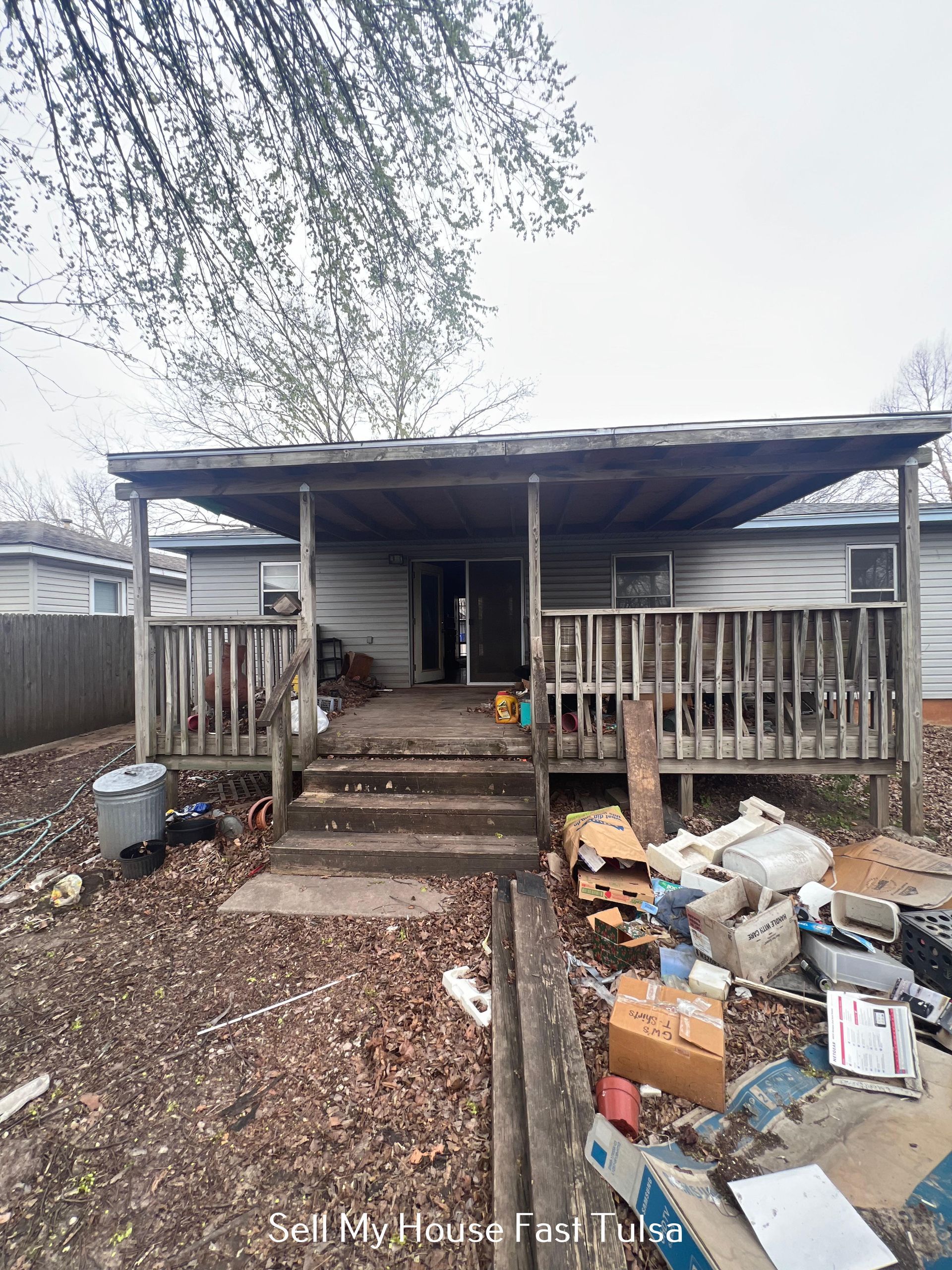 A weathered house with a wooden deck and porch, surrounded by scattered debris and trash in a backyard.