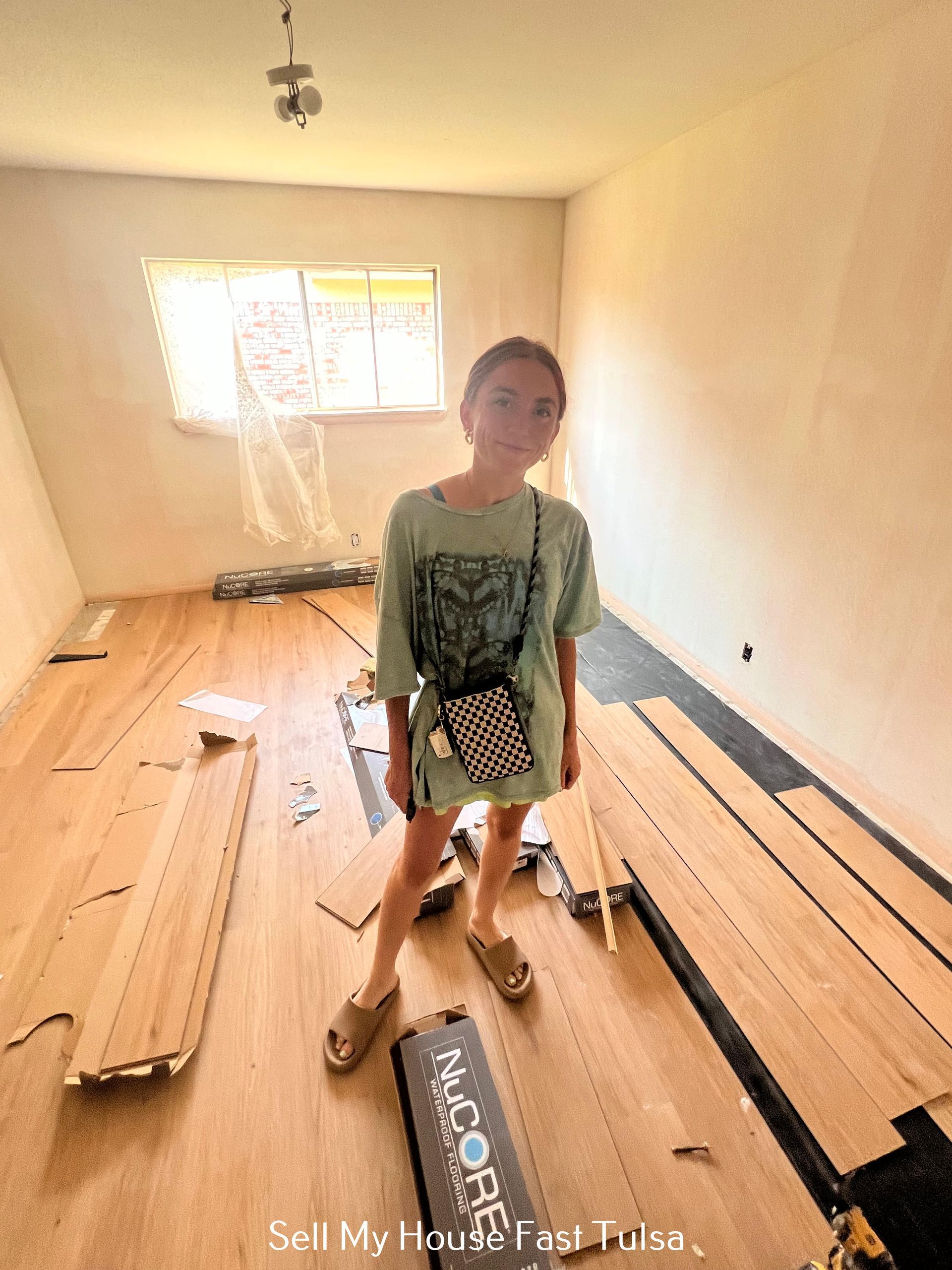 A person stands in a room under renovation with new wooden flooring boards scattered across the unfinished floor.