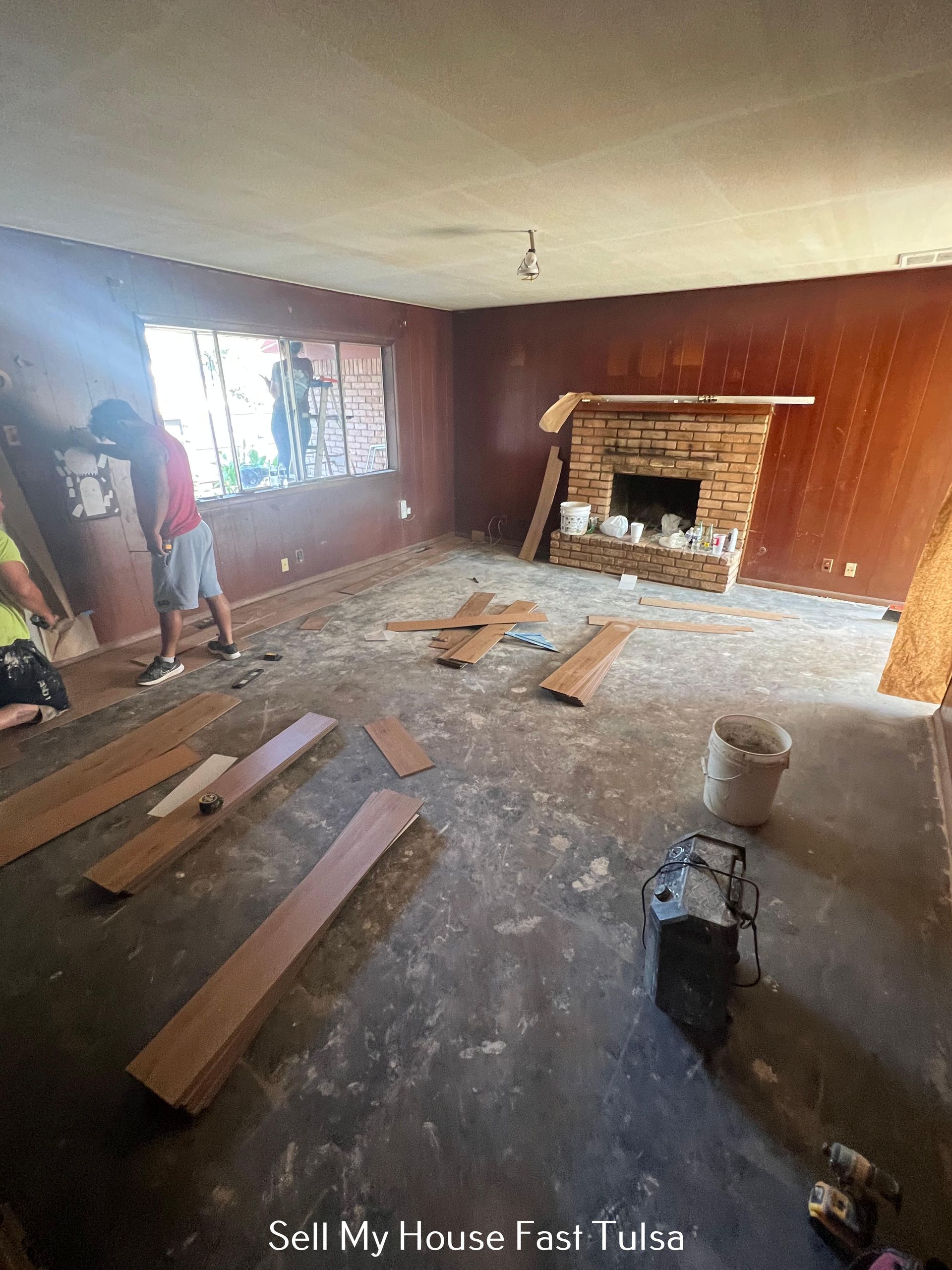 Living room under renovation with exposed floorboards, a fireplace, and a person by the window