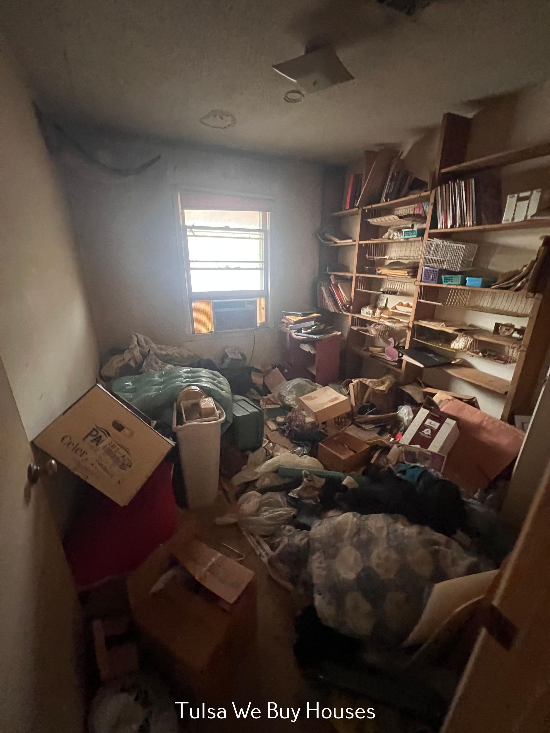 A cluttered, dim room filled with stacks of cardboard boxes, piles of personal items, and open shelving against the wall.