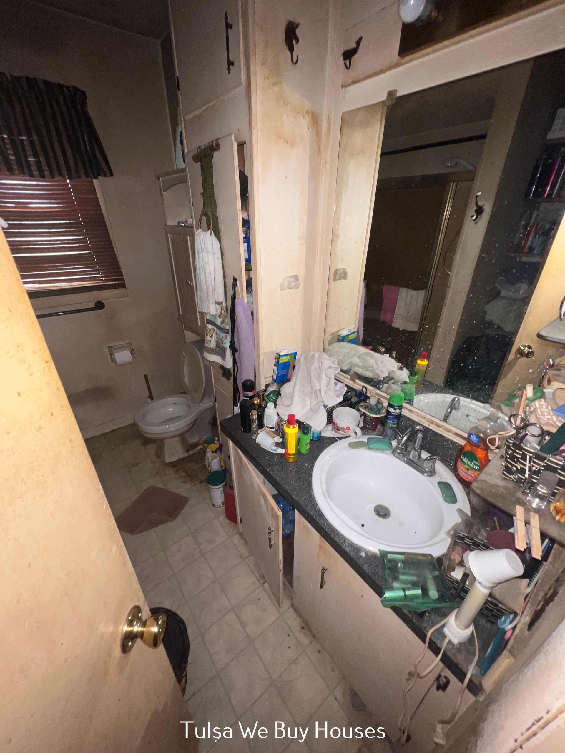 A cluttered bathroom featuring a white vanity with a sink, a toilet, a large mirror, and wood-paneled walls.