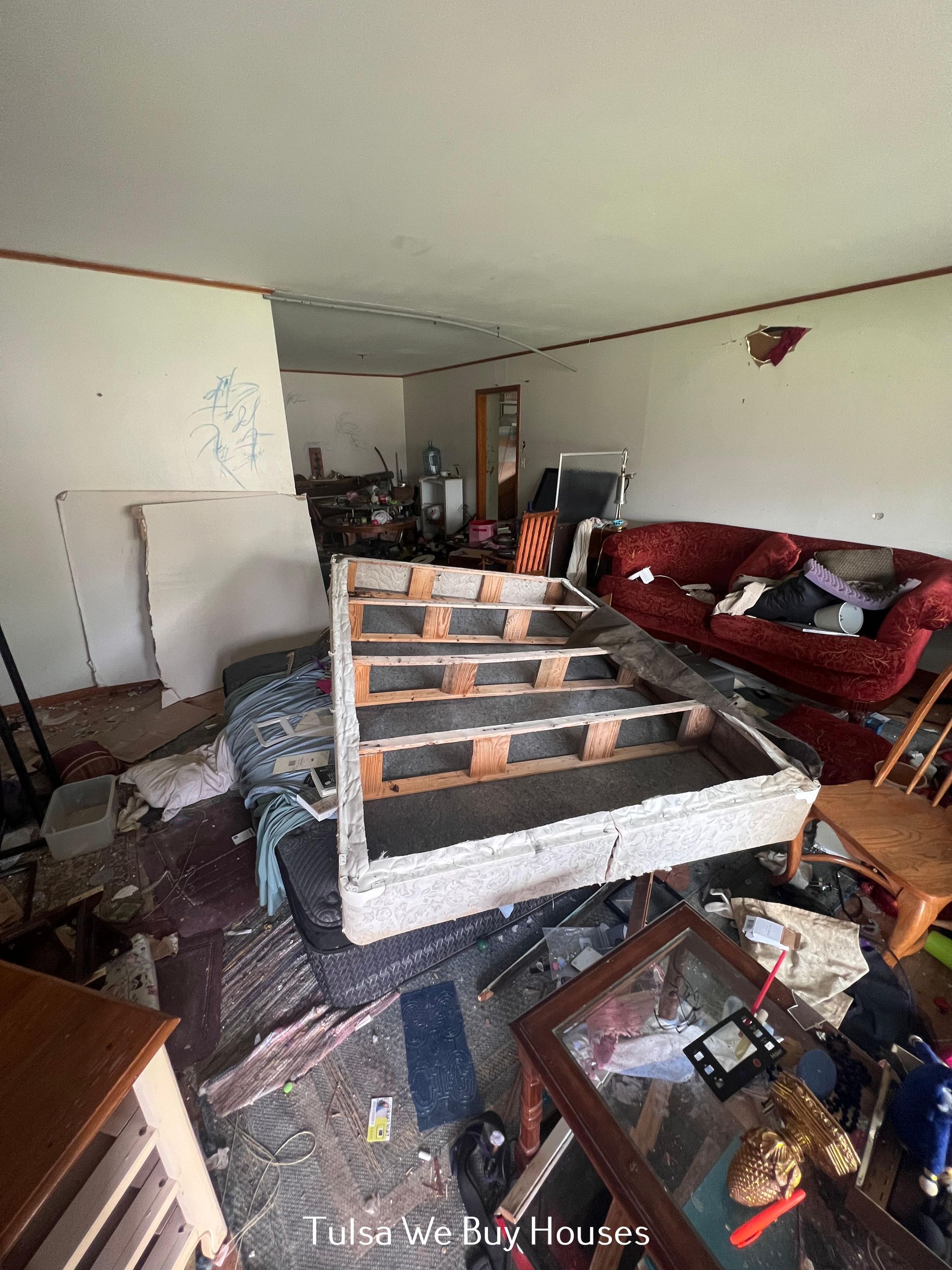 A cluttered living room with a bed frame in the center, a red couch, and debris scattered across the floor.