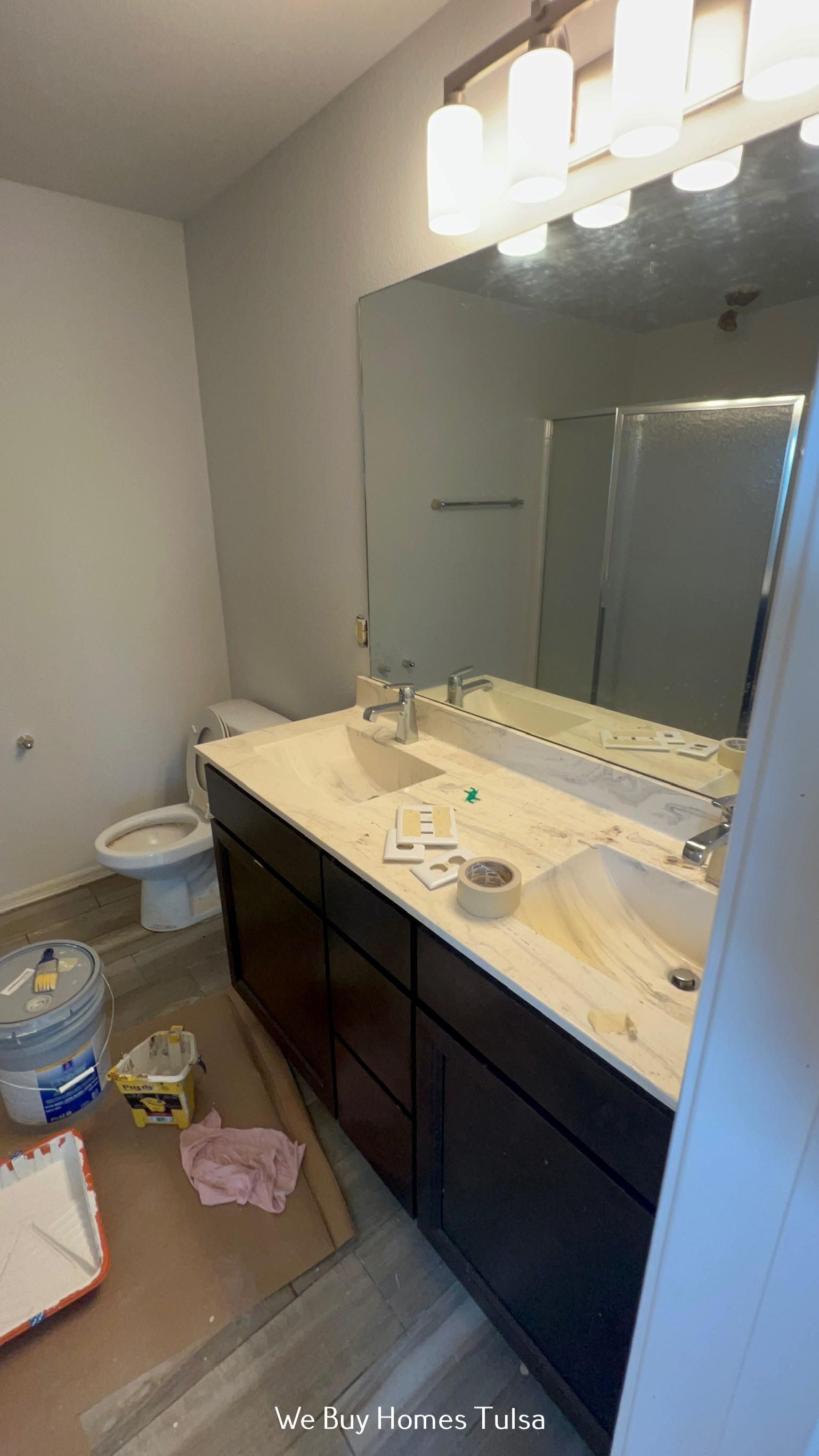A bathroom under construction with a dark wooden vanity, cream countertop, large mirror, and toilet, with paint supplies.