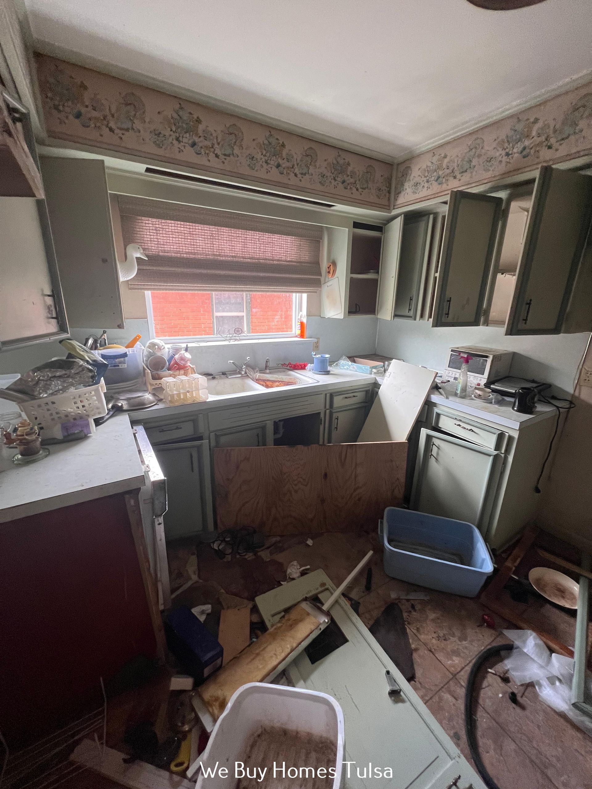 A cluttered kitchen with light green cabinets, debris on the floor, and a wooden panel blocking the sink area.