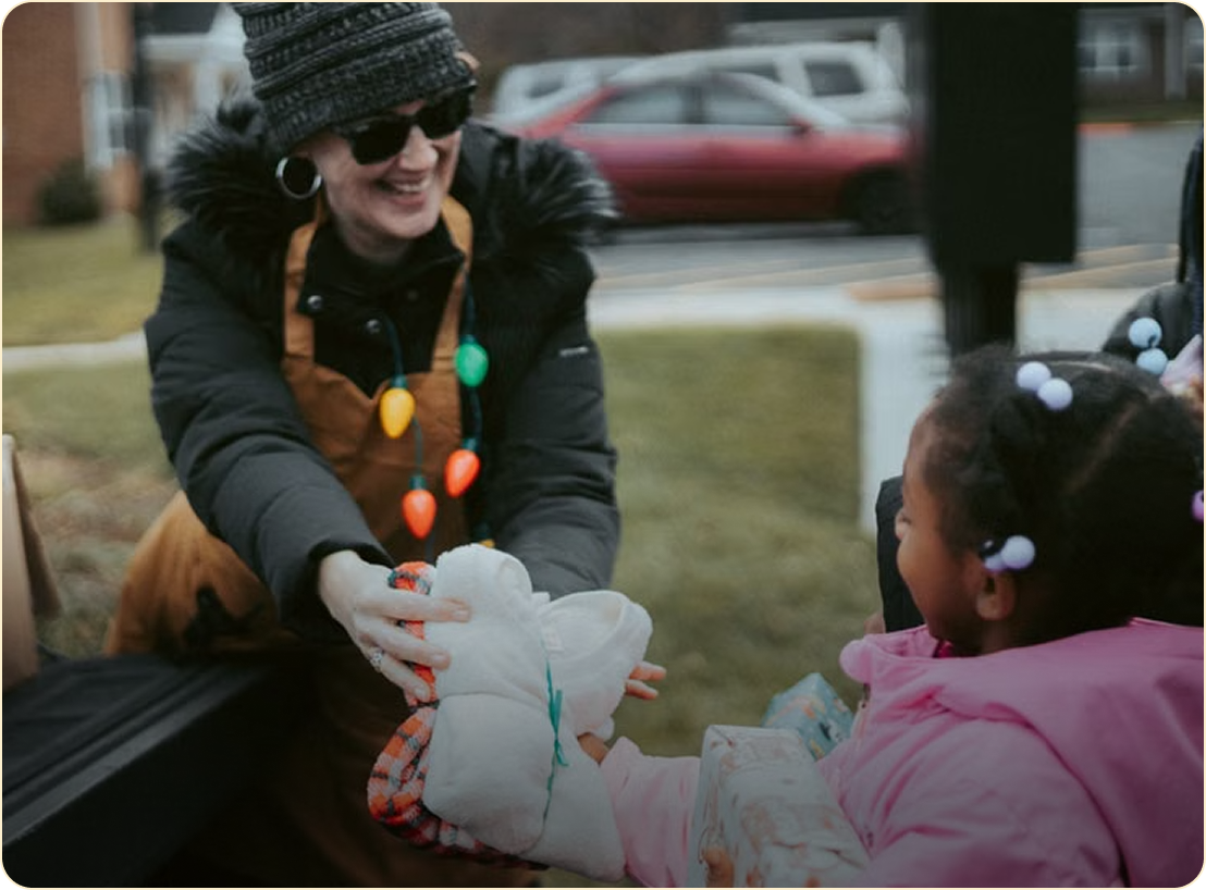 Woman in winter clothes passing a toy to a child outdoors, both smiling.