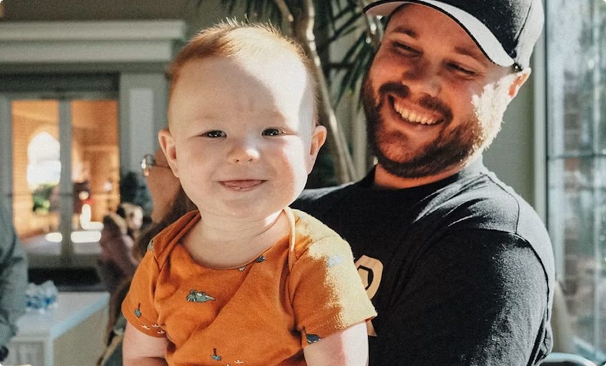 Smiling adult holding a toddler in an orange shirt indoors near a bright window