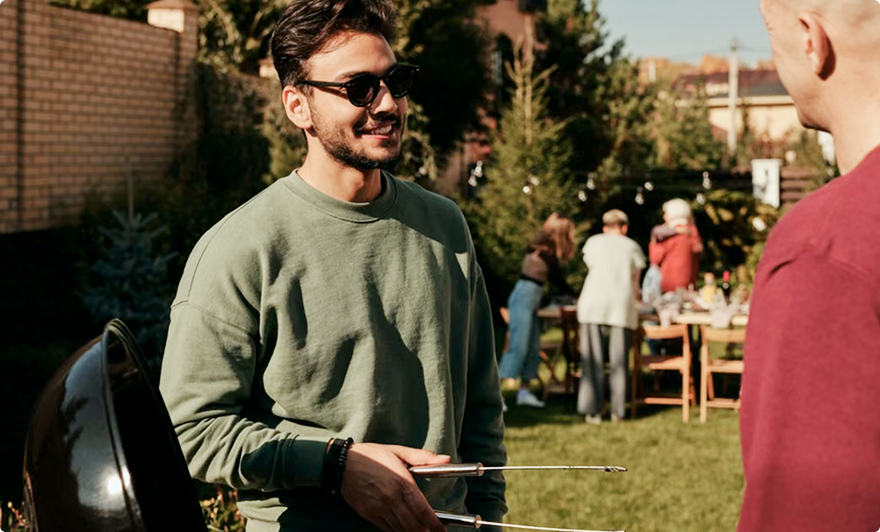 Man in sunglasses and green sweater talking outdoors at a garden gathering, holding a tray