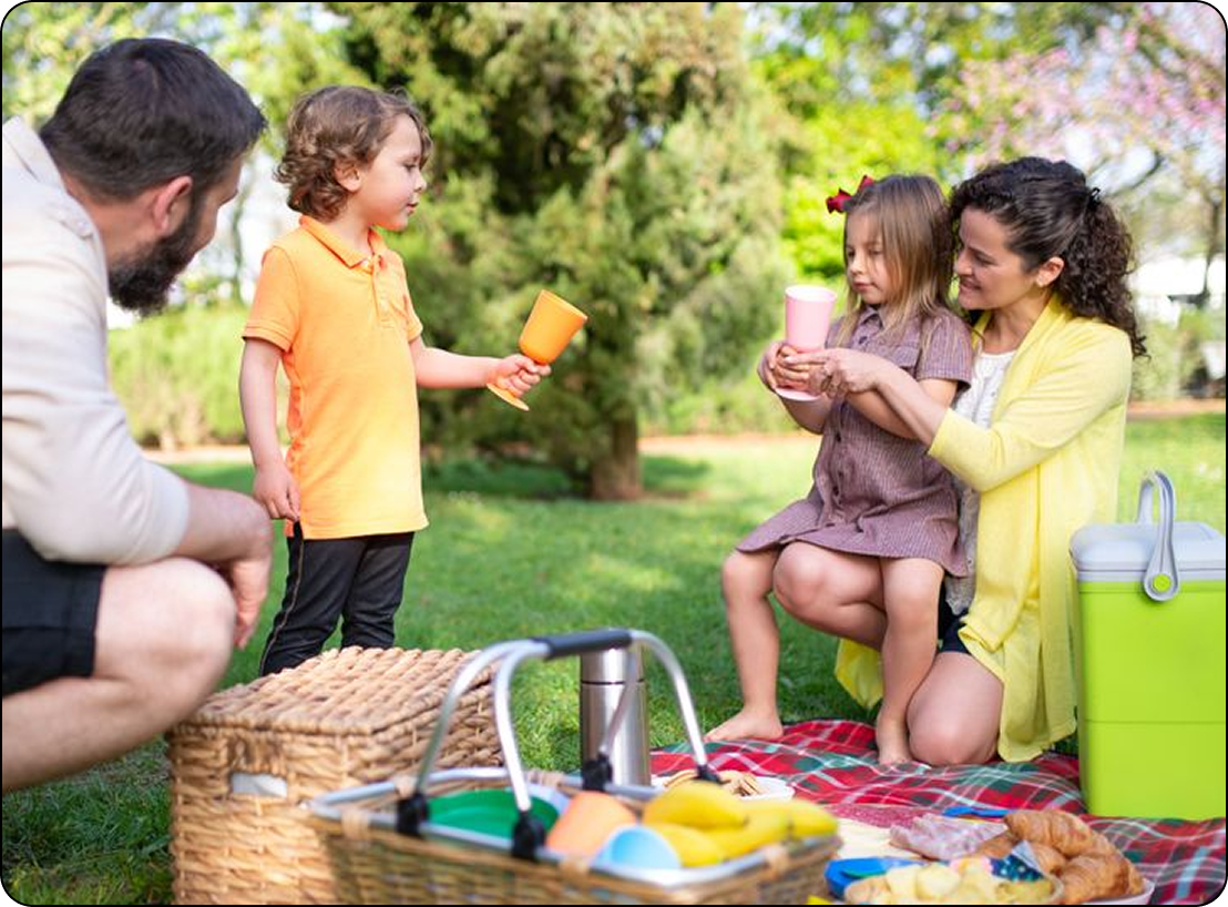 Family picnic in a park, with a child offering an orange cup to a seated adult and toddler.