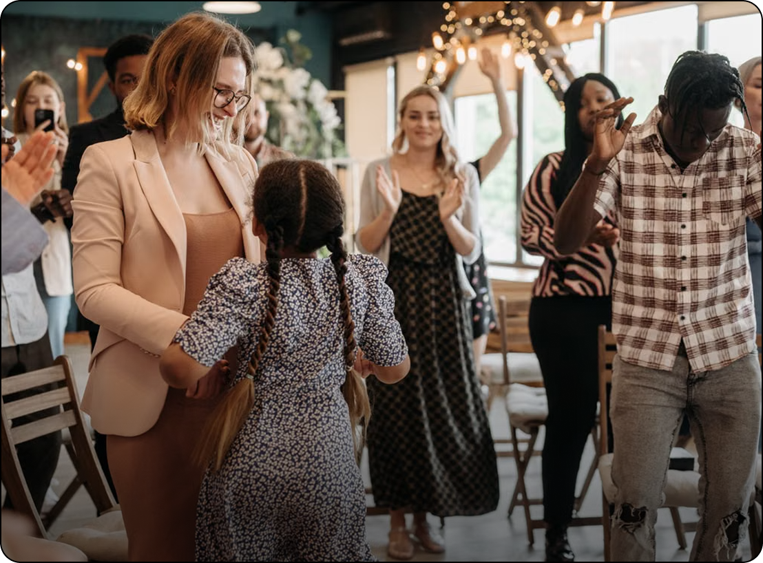 People socializing at a warmly lit indoor gathering, with a woman speaking to a child in the foreground