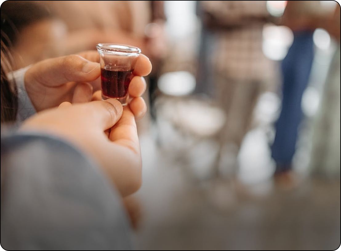 Hands holding a small glass of dark liquor in a blurred indoor setting