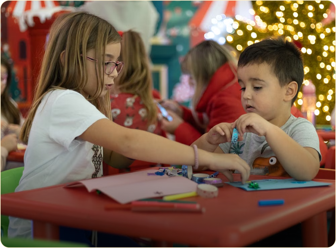 Children doing crafts at a red table, with colorful supplies and holiday lights in the background