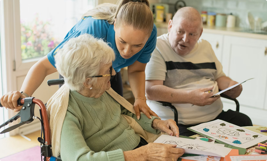 Caregiver helps an elderly woman with a craft activity while an older man watches nearby in a bright room