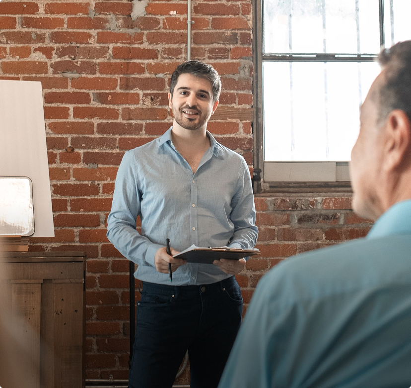 Man speaking to a group in a brick-walled office, holding a tablet near a bright window.