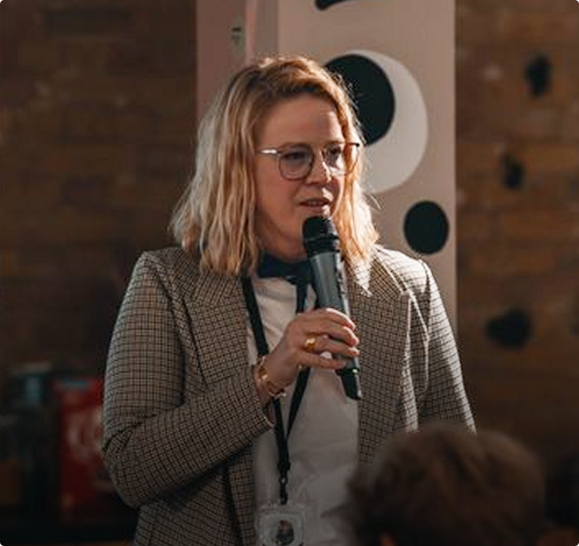 Speaker holding a microphone at an indoor event, wearing glasses and a checkered blazer.