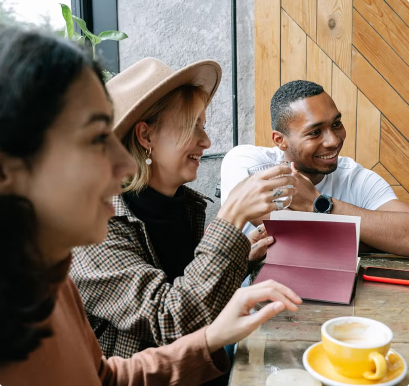 Friends chatting at a café table, smiling with drinks and a pink notebook.