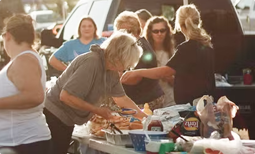 People serving food at an outdoor buffet beside parked cars in bright sunlight