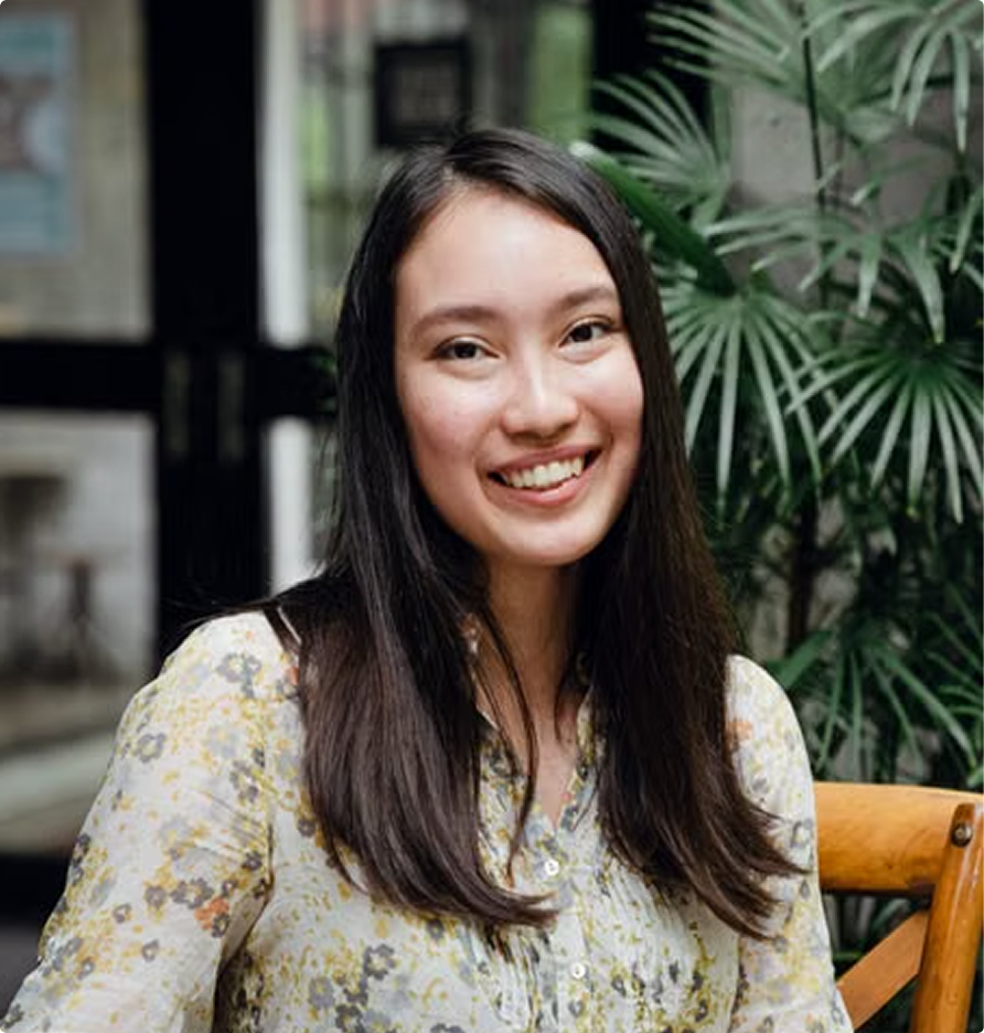 Smiling woman seated outdoors beside green plants, wearing a light floral blouse.