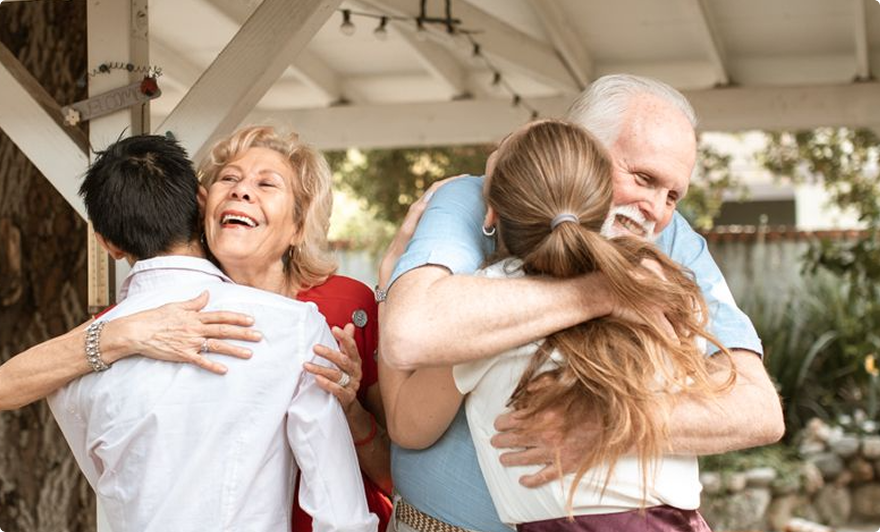 Two couples embracing in a bright indoor setting, smiling warmly and hugging each other.