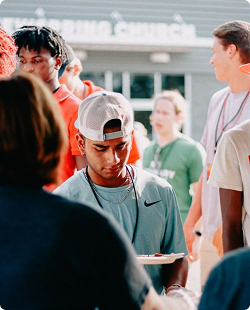 People gathered outdoors; a man in a gray cap looks down at a phone or paper.