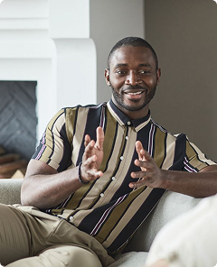 Smiling person in a striped shirt gesturing while seated on a couch in a living room