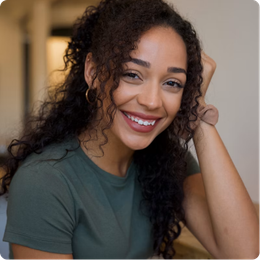 Smiling woman with curly hair in a green shirt, resting her head on her hand indoors.