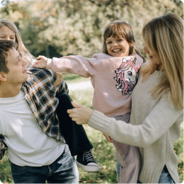 Family smiling and playing outdoors, with a child on an adult’s shoulders in a sunny park.
