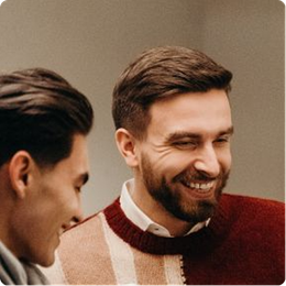 Two smiling men indoors, one in a red sweater, sharing a light moment.
