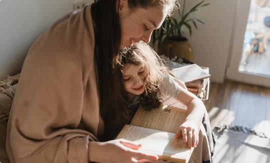 Woman and child reading a book together on a couch in warm sunlight