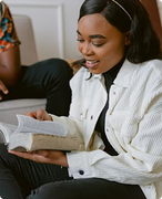 Woman in a white jacket reading a book and smiling on a couch
