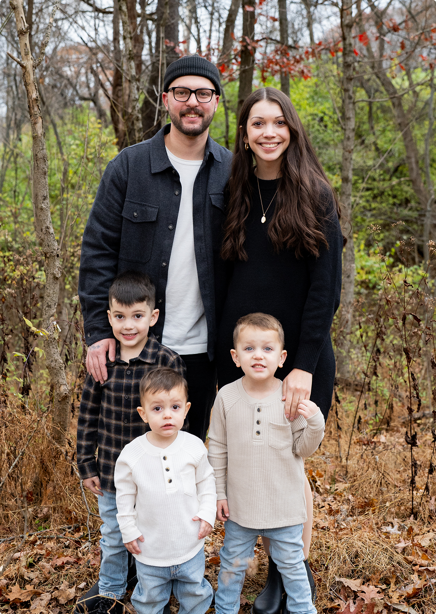 Family of five posing outdoors in a wooded area, smiling and standing close together.