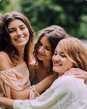Three smiling friends embracing outdoors in a green park, wearing light-colored tops.