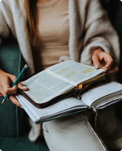 Person holding a pen over an open Bible and notebook, studying quietly indoors