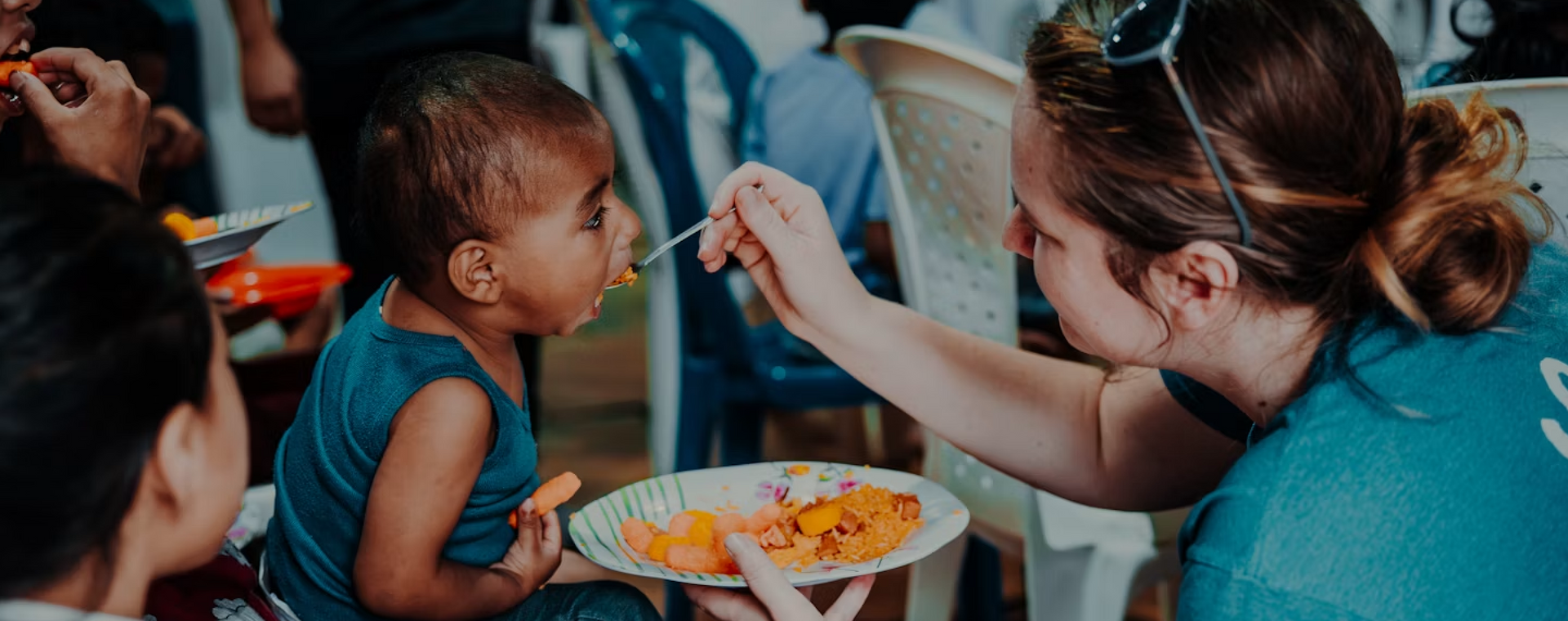 Woman feeding a toddler orange food at a table while others sit nearby
