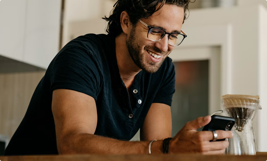 Smiling man in black shirt using a smartphone beside a pour-over coffee setup