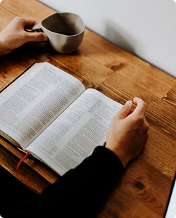 Hands holding an open book beside a mug on a wooden table