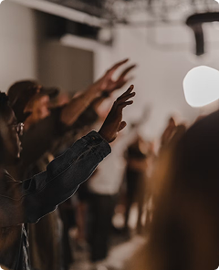 People raising hands in a dim indoor gathering, with a bright light in the background