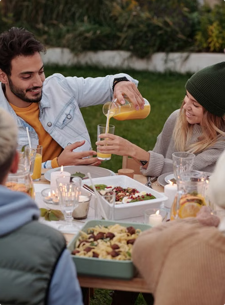 Friends sharing food and drinks at an outdoor dinner table in a garden, with candles lit