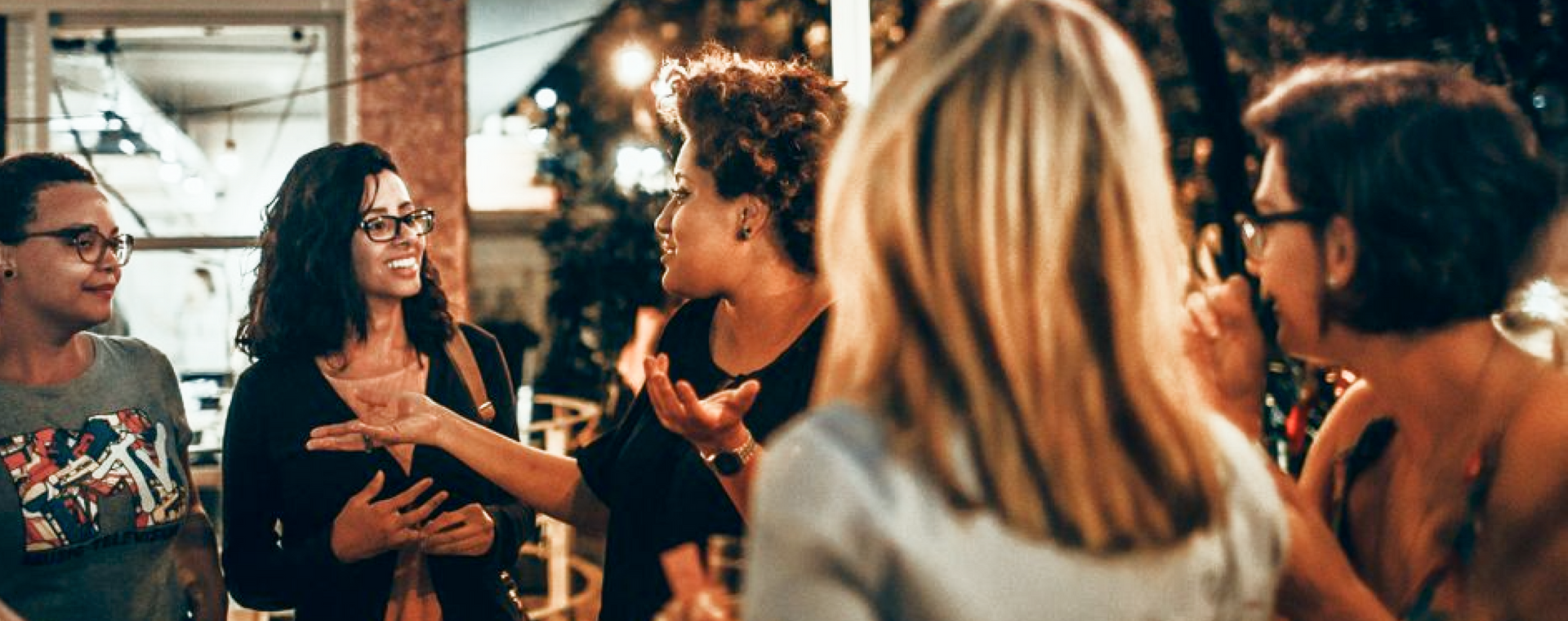People chatting at an indoor social gathering, with warm lights and blurred guests in the background